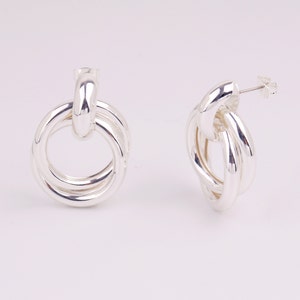 May include: A pair of silver hoop earrings with a knotted design. The earrings are made of a thick, smooth metal and have a polished finish.