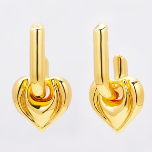 May include: A pair of gold-colored heart-shaped earrings. Each earring features a polished heart pendant connected to a vertical bar with a curved top. The earrings have a shiny, reflective surface.