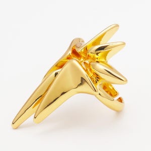 May include: A gold-colored ring with a starburst design. The ring features multiple pointed, triangular shapes radiating from a central point. The ring is made of a shiny, reflective metal.
