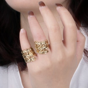 Könnte beinhalten: Zwei goldene, blattartige Ringe an einer Hand. Die Ringe sind breit und haben ein strukturiertes Design.