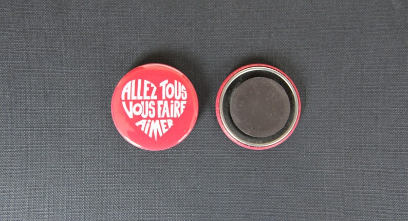 Puede incluir: Bot&oacute;n redondo rojo con un coraz&oacute;n blanco que contiene el texto "ALLEZ TOUS VOUS FAIRE AIMER" en franc&eacute;s. La parte posterior del bot&oacute;n es magn&eacute;tica.