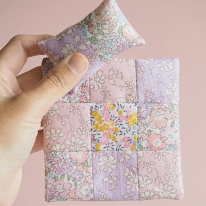 May include: A miniature patchwork pillow and coaster set. The floral fabric features pink, purple, and yellow hues. The pillow is triangular, and the coaster is square, both with a quilted design, perfect for a doll's house.