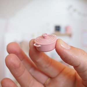 May include: A miniature pink cast iron pot with white handles. The pot is small enough to fit in the palm of a hand.