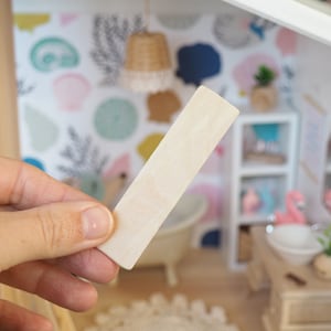 May include: A close-up of a plain, white wooden craft stick. The stick is being held by a hand in front of a dollhouse-sized bathroom with a bathtub, sink, and shelves.