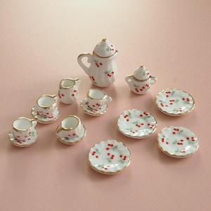 May include: A miniature white ceramic tea set with a cherry pattern. The set includes a teapot, sugar bowl, creamer, cups, saucers, and plates. Each piece has a gold trim and red cherry accents, ideal for a dollhouse or display.