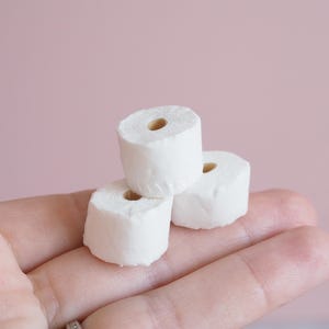 May include: Three miniature white toilet paper rolls stacked on a hand, with a light pink background. The rolls are small, with a visible cardboard core. The image is a close-up shot.