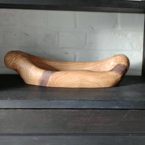 Peut inclure: Un bol en bois avec une finition en bois naturel. Le bol est en forme de banane et a une bande brun foncé autour du milieu.