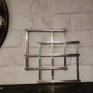 Peut inclure: Une étagère murale en métal argenté au design géométrique. L'étagère a une forme rectangulaire avec plusieurs barres horizontales et verticales, créant un motif en forme de grille. Elle est fixée sur un mur de briques blanches.