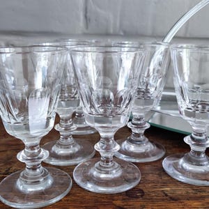May include: A set of six clear, stemmed glasses with a decorative etched pattern. The glasses have a rounded base and a detailed stem. They are arranged on a wooden surface, suggesting a vintage or antique aesthetic.