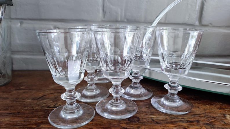 May include: A set of six clear, stemmed glasses with a decorative etched pattern. The glasses have a rounded base and a detailed stem. They are arranged on a wooden surface, suggesting a vintage or antique aesthetic.