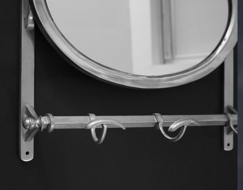 May include: A silver-toned wall-mounted mirror with a rounded oval shape and a decorative metal shelf with two hooks underneath.