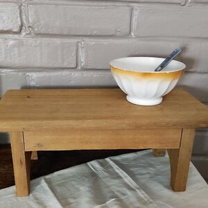 Peut inclure: Un petit tabouret rectangulaire en bois avec quatre pieds robustes et une finition lisse brun clair. Sur le dessus repose un bol en céramique blanche avec un bord festonné subtil et un rebord orange-brun délavé, contenant une cuillère en métal.