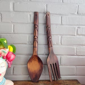 Puede incluir: Un juego de cuchara y tenedor de madera con diseños tallados de estilo tribal en los mangos. Los utensilios son de madera marrón oscuro y se exhiben contra una pared de ladrillos blancos. La cuchara tiene un cuenco redondeado y el tenedor tiene cuatro púas.