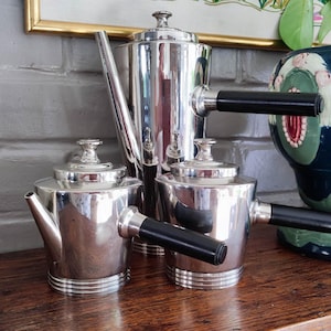 Könnte beinhalten: Ein silbernes Teeservice mit schwarzen Griffen und einer passenden Kaffeekanne. Das Set umfasst eine Teekanne, zwei kleine Töpfe und eine hohe Kaffeekanne. Ein dekorativer blau-grüner Keramiktopf ist ebenfalls sichtbar.