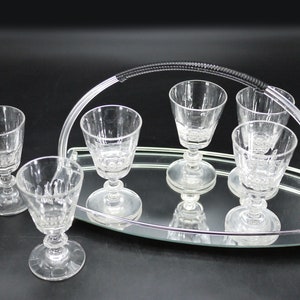 May include: A set of six clear glass cordial glasses on a mirrored tray with a silver handle. The glasses have a stemmed base and a patterned design. The tray is oval-shaped and reflects the glasses.