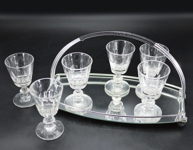 May include: A set of six clear glass cordial glasses on a mirrored tray with a silver handle. The glasses have a stemmed base and a patterned design. The tray is oval-shaped and reflects the glasses.