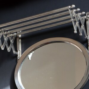 May include: A silver-toned, wall-mounted mirror with a fold-down clothes rack. The mirror is round and has a beveled edge. The rack has three bars and is attached to the mirror with a hinged arm.