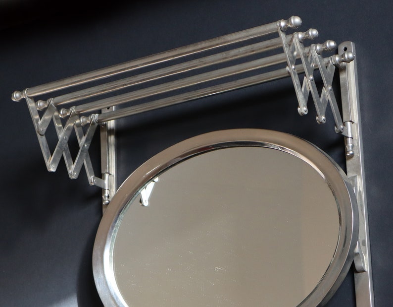 May include: A silver-toned, wall-mounted mirror with a fold-down clothes rack. The mirror is round and has a beveled edge. The rack has three bars and is attached to the mirror with a hinged arm.