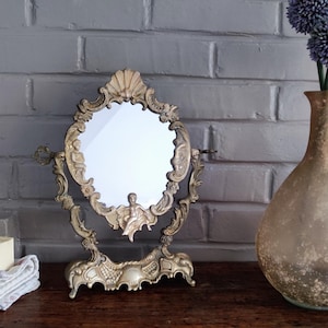 Peut inclure: Un miroir de coiffeuse de style antique avec un cadre doré orné. Le miroir ovale est soutenu par une base décorative et présente des motifs sculptés de chérubins et de coquillages. Le miroir est posé devant un mur de briques grises.