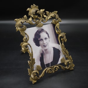 Peut inclure: Cadre photo doré orné avec une photographie vintage. Le cadre présente des motifs floraux et de volutes complexes. La photographie montre une femme en robe noire et collier de perles.