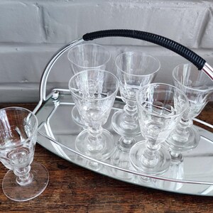 May include: A set of clear, stemmed cordial glasses on a silver-toned serving tray with a black handle. The glasses have a decorative pattern and are arranged neatly on the tray, perfect for serving drinks.
