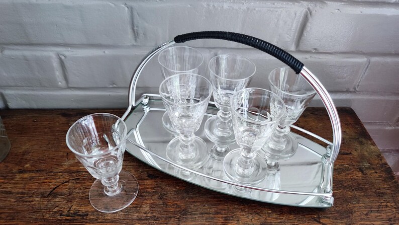 May include: A set of clear, stemmed cordial glasses on a silver-toned serving tray with a black handle. The glasses have a decorative pattern and are arranged neatly on the tray, perfect for serving drinks.