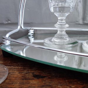 May include: Clear crystal wine glasses with ornate stems sit on a mirrored tray with a silver metal handle. The glasses reflect in the mirror, creating a symmetrical design. The tray is on a wooden surface.
