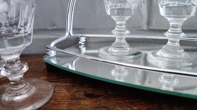 May include: Clear crystal wine glasses with ornate stems sit on a mirrored tray with a silver metal handle. The glasses reflect in the mirror, creating a symmetrical design. The tray is on a wooden surface.