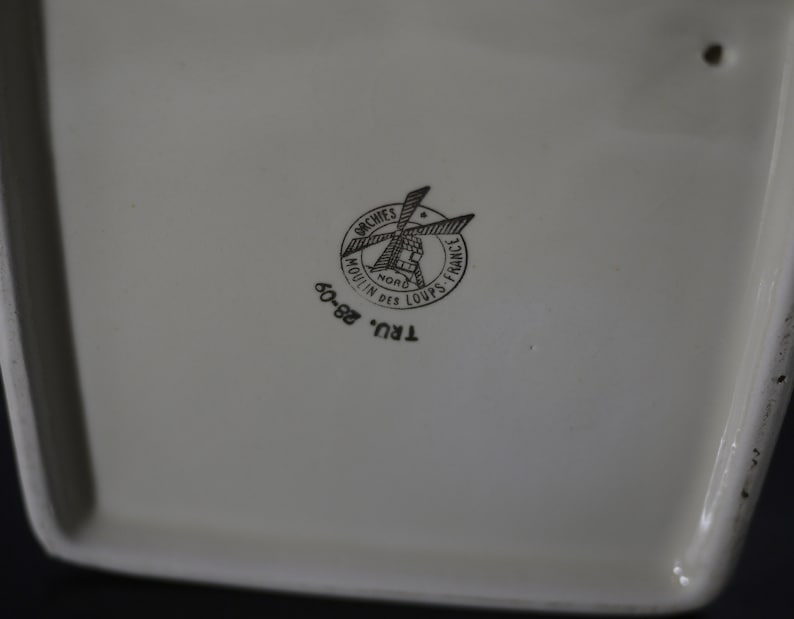 May include: Cream-colored ceramic piece with a black logo that reads "ORCHIES NORD MOULIN DES LOUPS FRANCE" and the numbers "60-82". The logo features a windmill design. The piece has a rectangular shape.