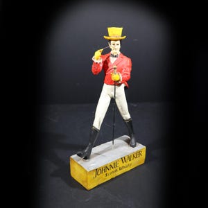 May include: Figurine of a man in a red jacket, white trousers, and black boots, holding a cane and a pipe. He wears a yellow top hat and gloves. The base reads "Johnnie Walker Scotch Whisky."