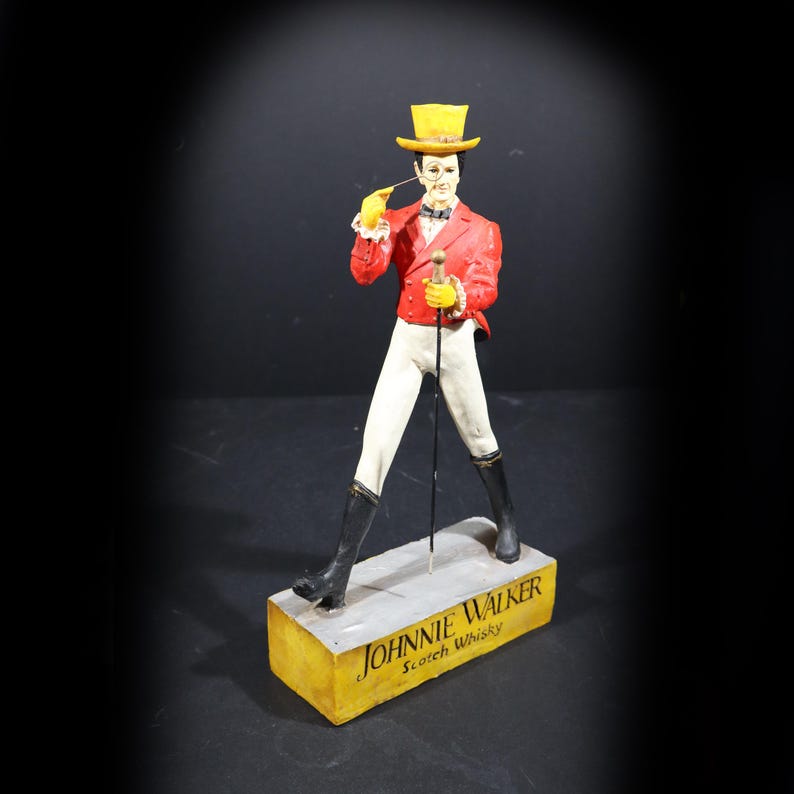 May include: Figurine of a man in a red jacket, white trousers, and black boots, holding a cane and a pipe. He wears a yellow top hat and gloves. The base reads "Johnnie Walker Scotch Whisky."