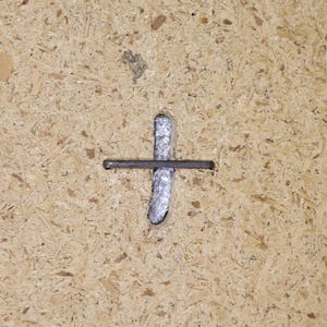 May include: A close-up of a metal nail hammered into a piece of particle board. The nail is bent at a 90-degree angle, creating a cross shape.