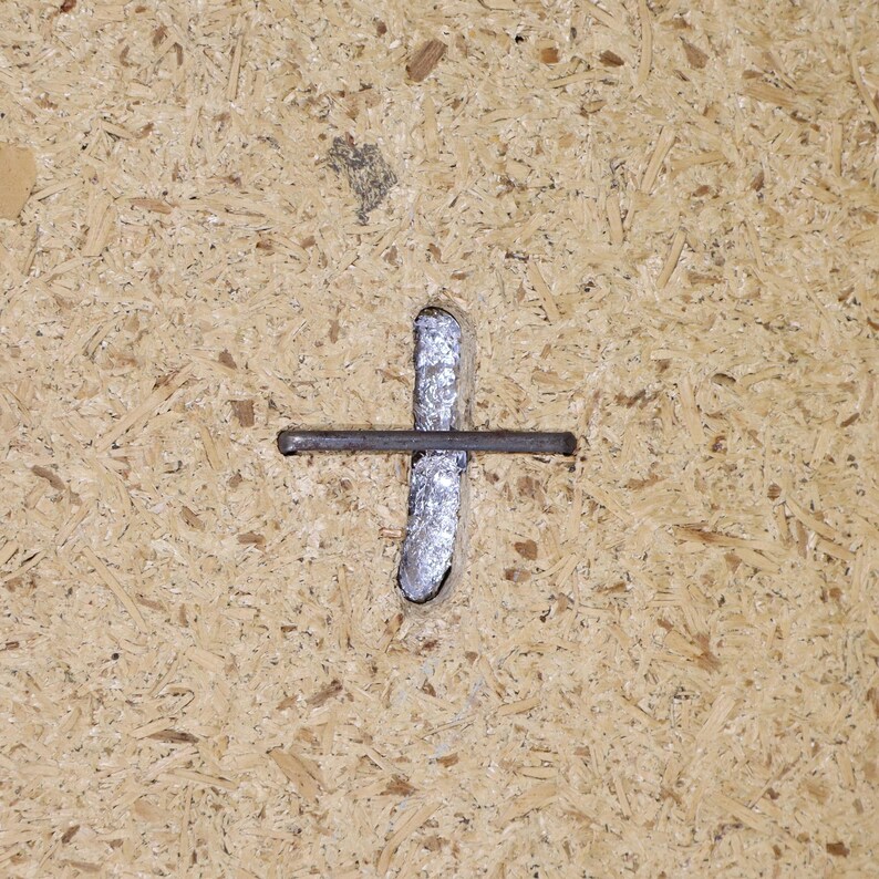 May include: A close-up of a metal nail hammered into a piece of particle board. The nail is bent at a 90-degree angle, creating a cross shape.