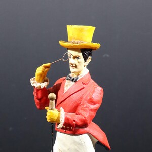 May include: A figurine of a man in a red tailcoat, yellow top hat, and monocle. The figure is holding a cane and wearing yellow gloves with white ruffled cuffs. The figure has a black bow tie and is set against a dark background.
