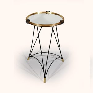 Peut inclure: Une table d'appoint ronde dotée d'un plateau miroir circulaire avec un cadre doré. La base se compose de fins pieds en métal noir qui s'entrecroisent selon un motif géométrique, se terminant par de petits pieds dorés à chaque extrémité.