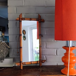 Peut inclure: Un miroir mural rectangulaire doté d'un cadre composé de segments de bambou polis avec des fixations brun foncé aux jointures. Le miroir est orienté verticalement et comporte une fine cordelette de suspension fixée sur le côté du cadre.