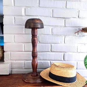 May include: A dark brown wooden hat stand with a smooth, rounded top, a turned pedestal base, and a central column. The stand features a visible wood grain pattern and a polished finish, positioned next to a straw hat with a black ribbon band.