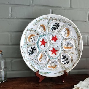 May include: A white ceramic decorative plate with a hand-painted design. The plate features red star shapes, brown and black abstract shapes, and a blue and green border. The plate is displayed on a wooden stand.