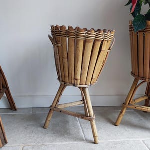 Peut inclure: Trois supports de plantes en bambou. Les supports sont de couleur bois naturel et présentent un design de panier tressé. Les supports sont soutenus par trois pieds. Un support est vide, et un autre contient une plante aux feuilles rouges.