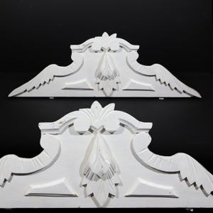 Può includere: Due elementi architettonici in legno bianco ornati. Ogni pezzo presenta un motivo floreale centrale con foglie stilizzate ed estensioni simmetriche a forma di ala. I pezzi sono dipinti di bianco e hanno un aspetto vintage invecchiato.