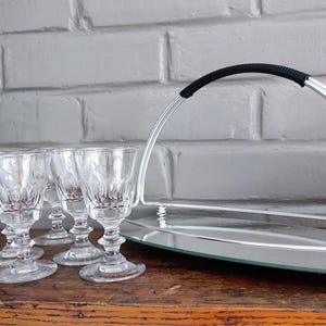 May include: A set of six clear, stemmed glasses with a decorative pattern, arranged on a wooden surface. A silver serving tray with a black handle and a mirrored base is also present, along with an ice bucket.