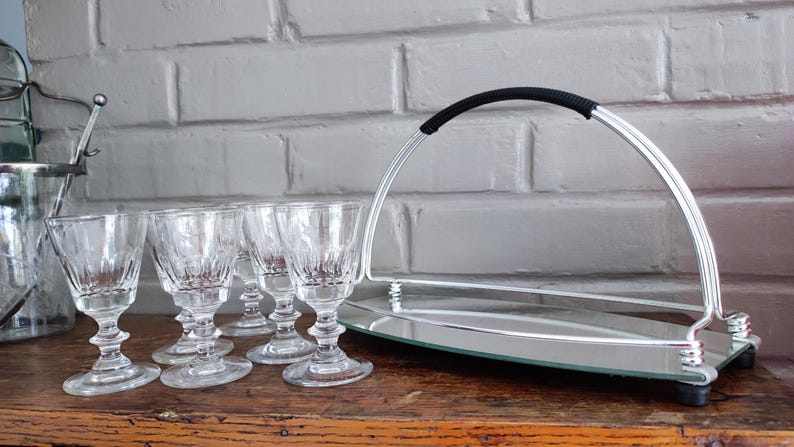 May include: A set of six clear, stemmed glasses with a decorative pattern, arranged on a wooden surface. A silver serving tray with a black handle and a mirrored base is also present, along with an ice bucket.