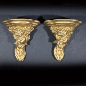 May include: A pair of gold-painted wall shelf brackets with a cherub design. Each bracket features a cherub holding a flower and a leafy design.