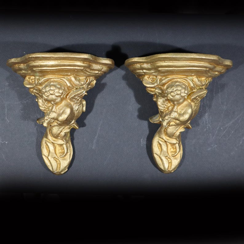 May include: A pair of gold-painted wall shelf brackets with a cherub design. Each bracket features a cherub holding a flower and a leafy design.