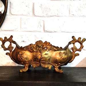 Puede incluir: Un cuenco central alargado de metal dorado con motivos ornamentales en relieve de flores y hojas. El recipiente cuenta con dos asas decorativas caladas a cada lado y se apoya sobre cuatro pequeñas patas curvas, con un acabado desgastado.