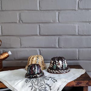 Puede incluir: Tres moldes de pudín de cerámica con diseños florales y de remolinos. Los moldes son en tonos marrón, crema y negro, y se asientan sobre un paño blanco con un patrón decorativo. Los moldes están sobre una mesa de madera contra una pared de ladrillo gris.