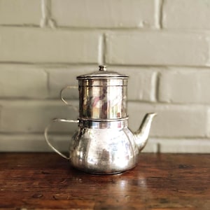 Peut inclure: Une cafetière en métal argenté dotée d'une base arrondie, d'un bec verseur incurvé et d'une poignée latérale. La partie supérieure comprend une chambre de filtration cylindrique surmontée d'un petit couvercle et d'une seconde poignée.