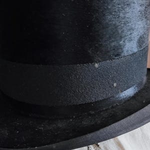 May include: A black top hat featuring a tall, cylindrical crown and a wide, curved brim. The hat is encircled by a textured black grosgrain ribbon band that sits just above the brim, providing a distinct contrast to the smooth finish of the hat.