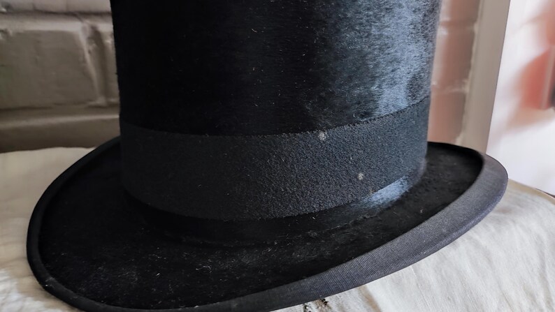 May include: A black top hat featuring a tall, cylindrical crown and a wide, curved brim. The hat is encircled by a textured black grosgrain ribbon band that sits just above the brim, providing a distinct contrast to the smooth finish of the hat.