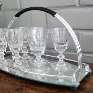 May include: A silver serving tray with six clear, stemmed glasses. The tray has a mirrored surface and a curved handle with a black grip. An ice bucket is visible in the background. The glasses are approximately 4 inches tall.
